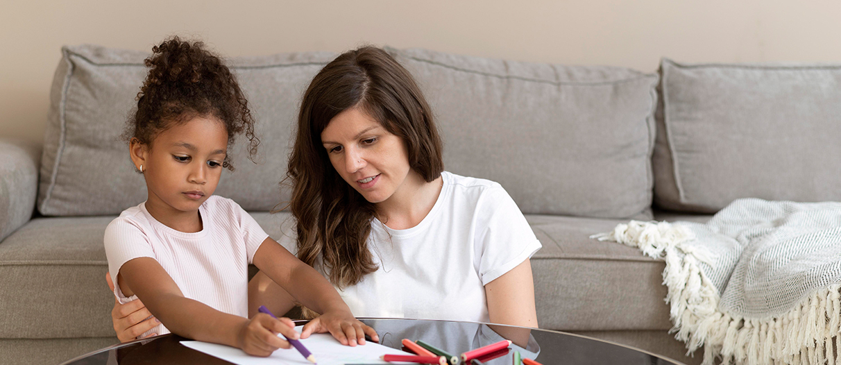 A girl child with her mother doing sketch work on the glass table.